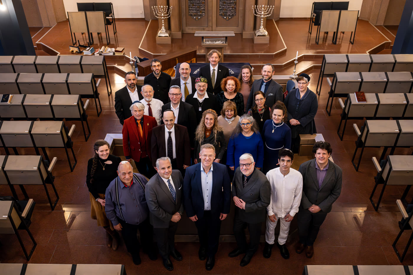 Some two dozen cantors gathered for a group photograph in the Mannheim synagogue, seen from above, with the ark, two silver menorahs and the empty desks of the sanctuary behind them.