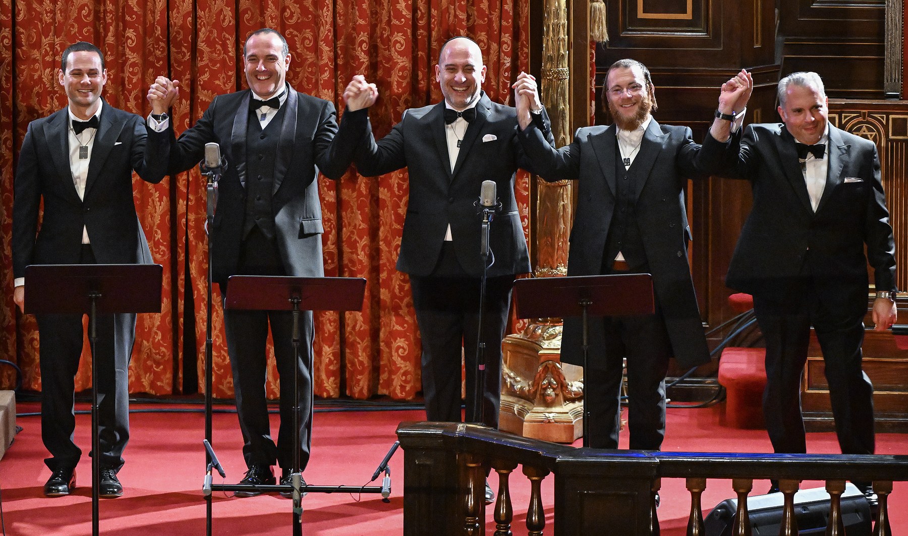 Five cantors in black tie standing in a line before the red curtain of the ark, hands joined and raised above their heads, laughing, their music stands in front of them.