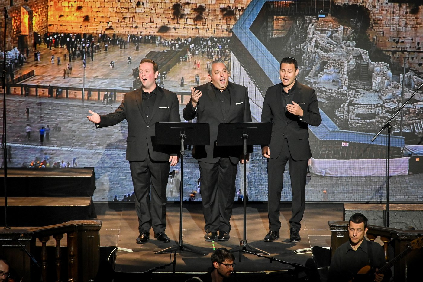 Gideon Zelermyer, Chaim David Berson and Azi Schwartz singing at music stands in front of a full-height image of the Western Wall plaza.