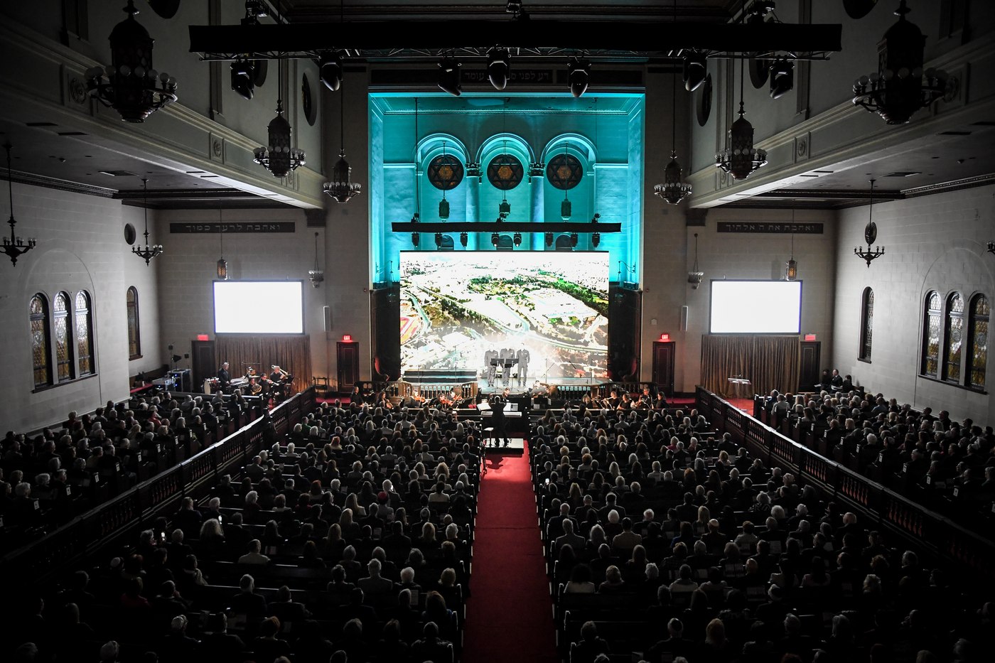 The sanctuary from the balcony, every pew full, the three cantors small on a platform in front of a vast lit screen showing an aerial view of Jerusalem, the orchestra below them.