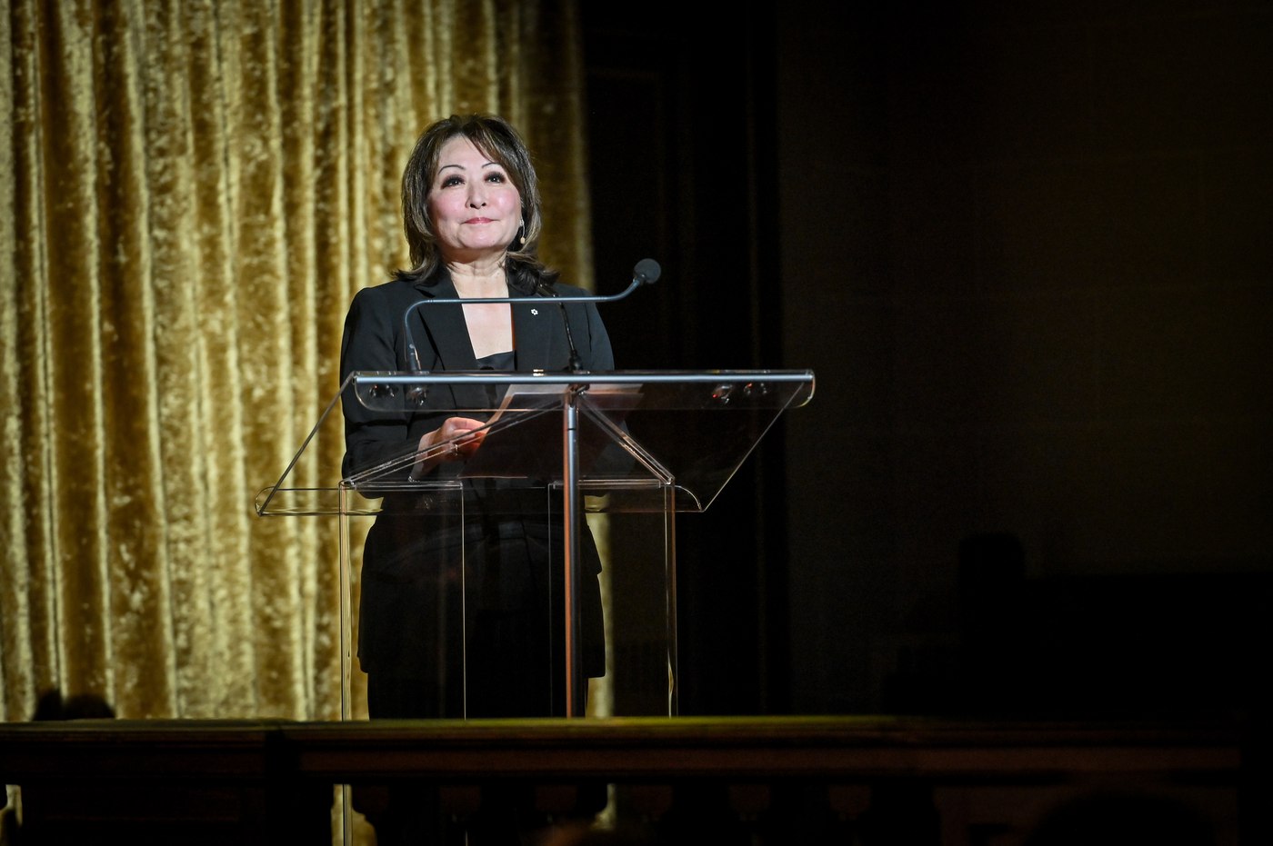 Mutsumi Takahashi at a glass lectern in a spotlight, the gold stage curtain behind her.