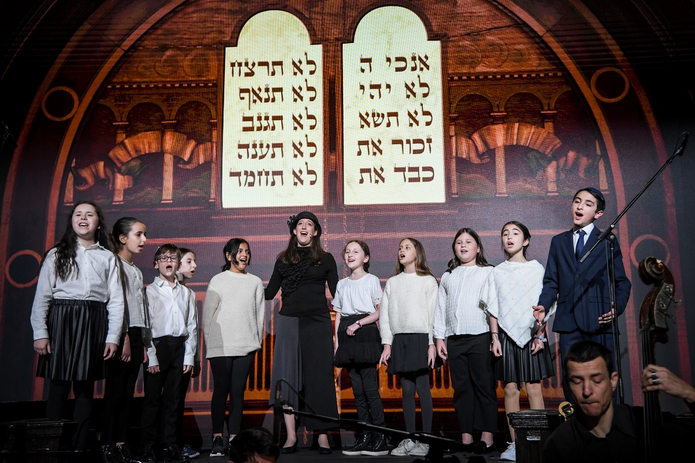 Rabba Rachel Kohl-Finegold in a black hat singing with ten children of the Shaar Akiva Youth Choir, the Ten Commandments projected on the wall behind them.
